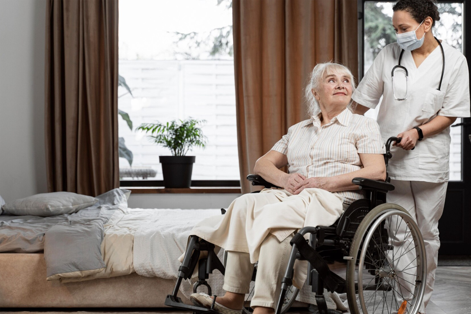 Caregiver in scrubs assisting an elderly resident in a wheelchair in a care-home room